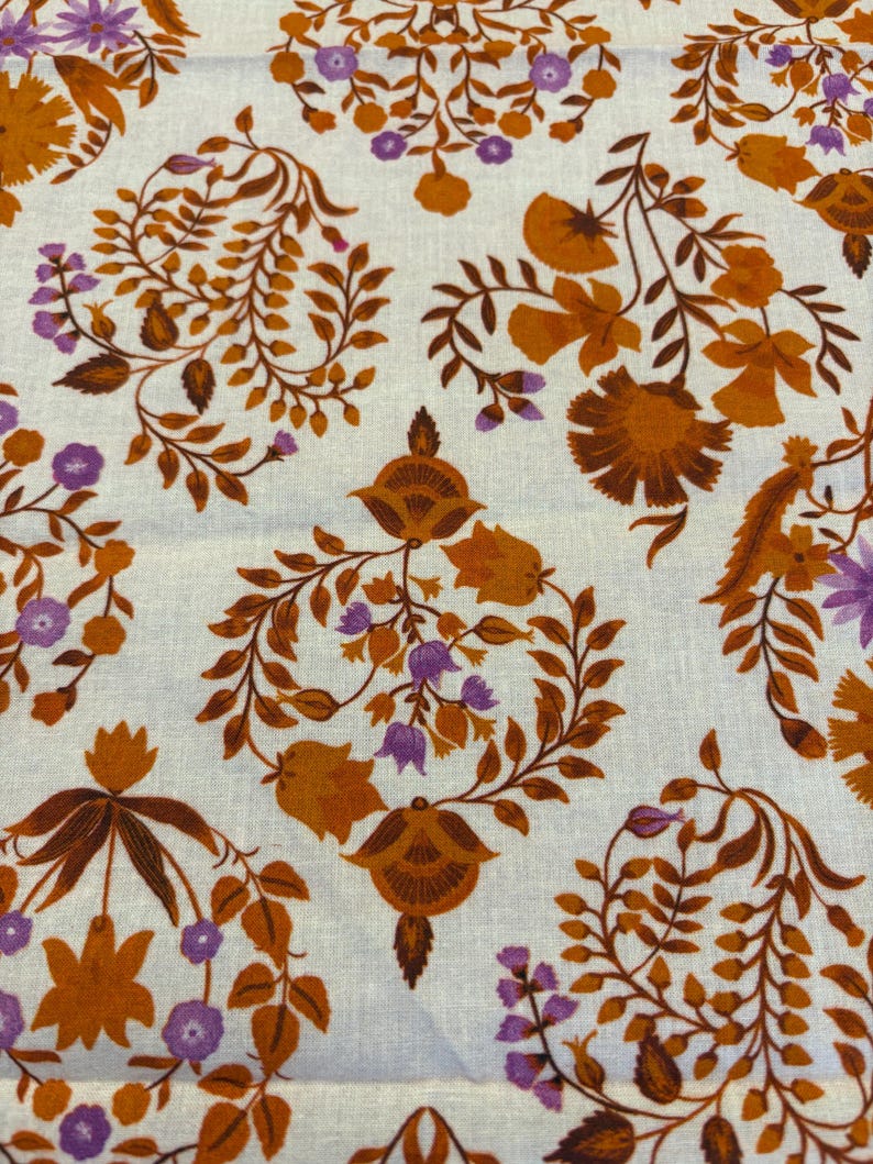 Op de afbeelding: Close-up van een stof met een herhalend bloemenpatroon. Het ontwerp toont gestileerde bloemen en bladeren in tinten oranje, bruin en paars tegen een cr&egrave;mekleurige achtergrond. Het patroon is symmetrisch en gedetailleerd, wat een vintage esthetiek cre&euml;ert.
