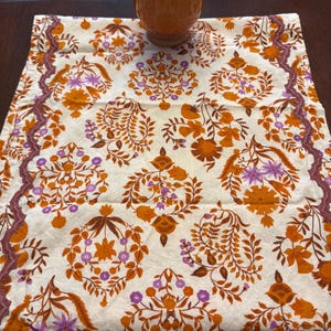 Op de afbeelding: Een decoratieve tafelloper met een bloemenpatroon in oranje, paars en bruin op een cr&egrave;mekleurige achtergrond. Een oranje kaars staat op de loper. De randen van de loper hebben een decoratieve rand.