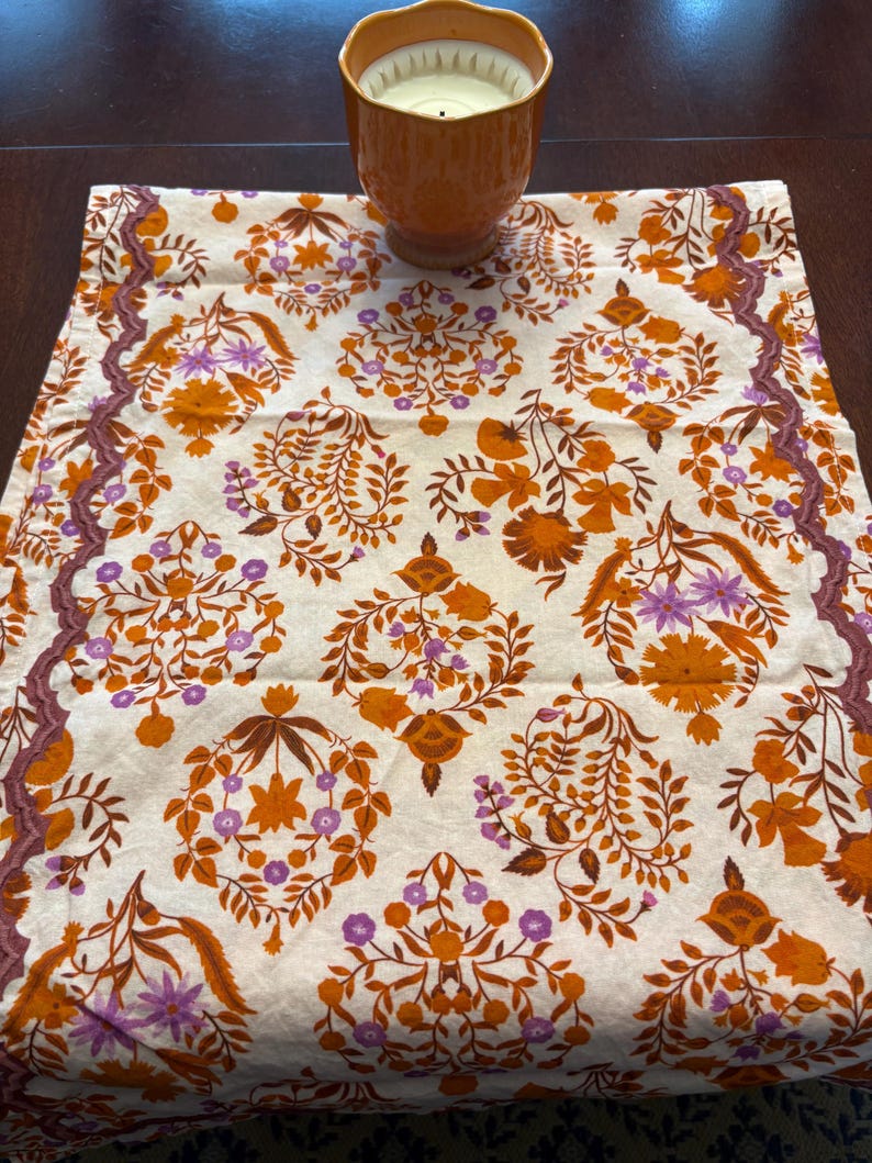 Op de afbeelding: Een decoratieve tafelloper met een bloemenpatroon in oranje, paars en bruin op een cr&egrave;mekleurige achtergrond. Een oranje kaars staat op de loper. De randen van de loper hebben een decoratieve rand.