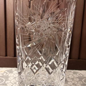 May include: A clear, rectangular crystal vase with intricate starburst and diamond-cut patterns. The vase has a textured surface and a decorative rim. This elegant piece is suitable for displaying flowers or as a decorative accent.