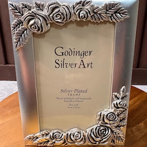 May include: A silver-plated picture frame featuring embossed rose and leaf motifs at the top left and bottom right corners. The insert displays the text Godinger Silver Art, Silver-Plated FRAME, Hand polished and lacquered, tarnish-resistant, 3 1/2" x 5".