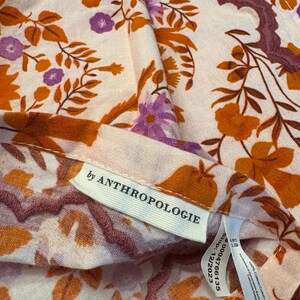 Op de afbeelding: Close-up van een stof met bloemenpatroon met een cr&egrave;mekleurige achtergrond. Het ontwerp bevat oranje en paarse bloemen en bladeren. Het label luidt "by ANTHROPOLOGIE".