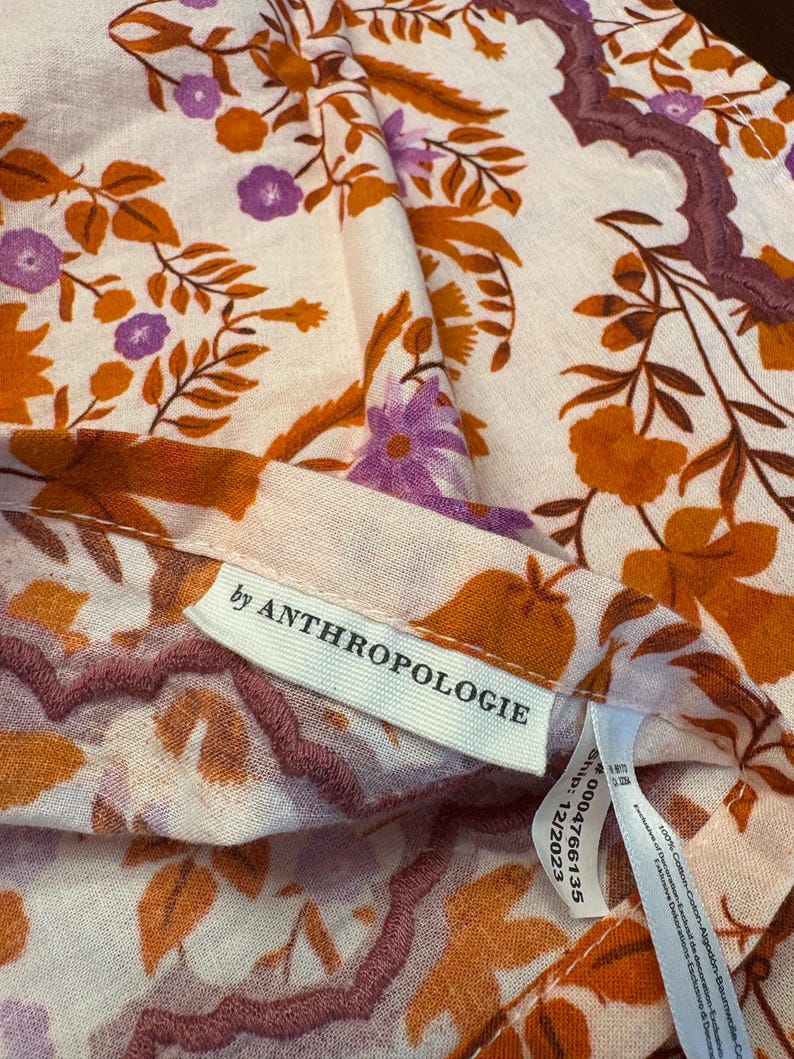 Op de afbeelding: Close-up van een stof met bloemenpatroon met een cr&egrave;mekleurige achtergrond. Het ontwerp bevat oranje en paarse bloemen en bladeren. Het label luidt "by ANTHROPOLOGIE".