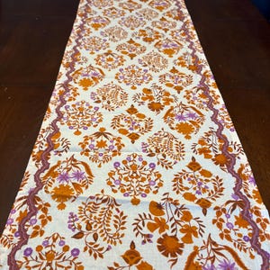 Op de afbeelding: Cr&egrave;mekleurige tafelloper met een herhalend bloemenpatroon in oranje en paarse tinten. De loper heeft een decoratieve rand in een donkerdere paarse tint. Het bloemmotief omvat gestileerde bloemen en bladeren.