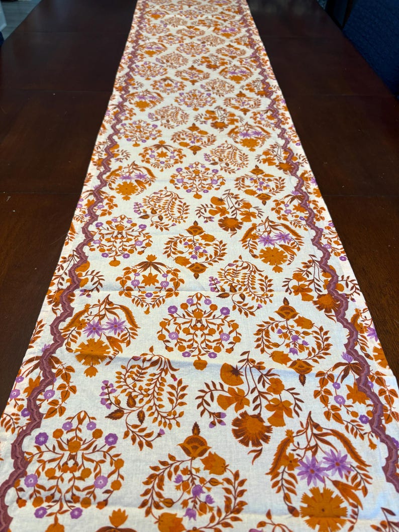 Op de afbeelding: Cr&egrave;mekleurige tafelloper met een herhalend bloemenpatroon in oranje en paarse tinten. De loper heeft een decoratieve rand in een donkerdere paarse tint. Het bloemmotief omvat gestileerde bloemen en bladeren.