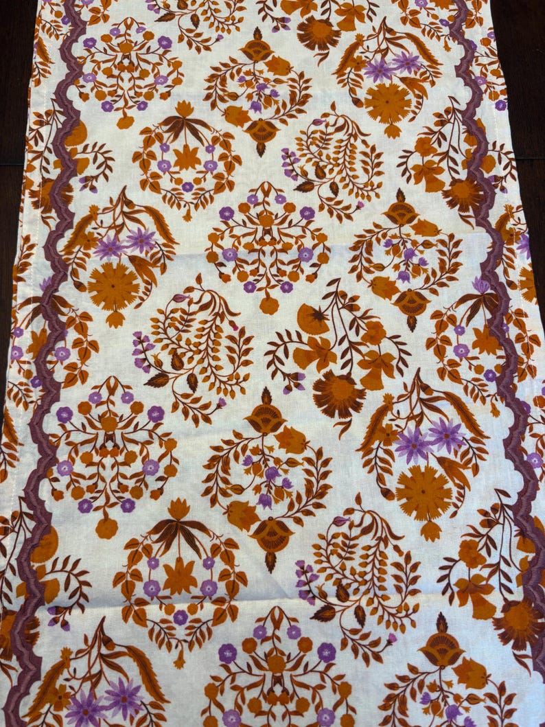 Op de afbeelding: Een witte tafelloper met een herhalend bloemenpatroon in tinten oranje, bruin en paars. Het ontwerp bevat gestileerde bloemen en bladeren. Een donkerpaarse rand met een rankachtig patroon loopt langs de randen.