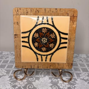 May include: A square cork-framed tile holder with a decorative ceramic tile. The tile features a floral design in brown, black, and cream colors. The holder has two metal hooks on the bottom.