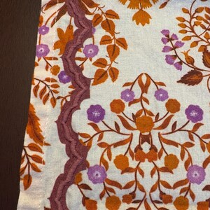 Op de afbeelding: Close-up van een stof met een bloemenpatroon. Het ontwerp toont oranje, paarse en bruine bloemen en bladeren op een lichte achtergrond. Een decoratieve, geschulpte rand in een dieppaarse kleur loopt langs de rand van de stof.