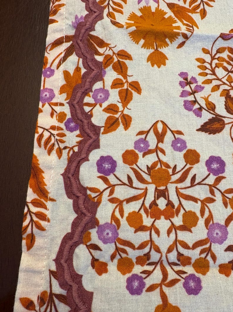 Op de afbeelding: Close-up van een stof met een bloemenpatroon. Het ontwerp toont oranje, paarse en bruine bloemen en bladeren op een lichte achtergrond. Een decoratieve, geschulpte rand in een dieppaarse kleur loopt langs de rand van de stof.