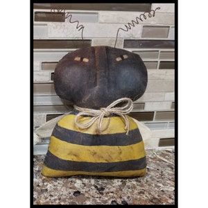 May include: A handmade black and yellow bee doll with a brown face and wire antennae. The bee is wearing a brown and yellow striped body and has a brown fabric bow tied around its neck.