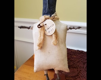 May include: A handmade rag doll with a black fabric head, white fabric body, and twig arms and legs. The doll is wearing a tan fabric scarf and has a safety pin attached to the scarf. The doll is standing on a wooden base and has a tag that says "Prairie Doll".