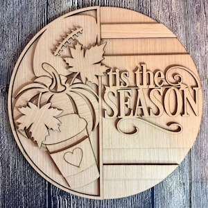 May include: A wooden round sign with a fall theme. The sign features a pumpkin, leaves, and a coffee cup with a heart. The words "tis the SEASON" are cut out of the wood. The sign is unfinished and ready to paint.