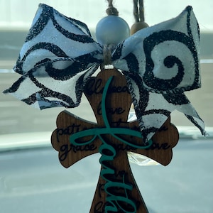 May include: A hanging wooden cross ornament featuring the word faith in teal script layered over a brown wood background with engraved words including blessed, love, grace, and patience. A white and black glittery bow sits at the top.