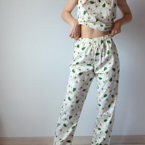 May include: White pajama set with a repeating avocado pattern. The set includes a camisole top and loose-fitting pants. The avocados are green with brown seeds and are scattered across the fabric. The pajamas are made of a lightweight material.