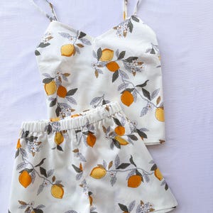Könnte beinhalten: Ein weißes Pyjama-Set mit Zitronenmuster. Das Set besteht aus einem Camisole-Top mit dünnen Trägern und passenden Shorts. Der Stoff ist mit gelben Zitronen, orangen Früchten, grünen Blättern und weißen Blüten gemustert.