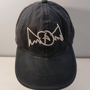 May include: A black baseball cap featuring an embroidered white graphic on the front panel. The design consists of a circular emblem with a central letter A, flanked by two stylized wing shapes. The cap has a curved brim and a button on the crown.