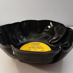May include: A black, upcycled vinyl record bowl with a wavy, scalloped edge. The yellow record label in the center reads "Tammy Wynette's Greatest Hits."