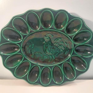 May include: Green ceramic egg serving platter with a scalloped edge. The center features a raised relief of a rooster. The platter has 12 egg-shaped indentations around the edge, each approximately 2.5 inches long.