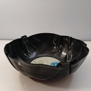 May include: A black bowl crafted from a repurposed vinyl record with a wavy, folded rim. The center of the bowl features a circular white label with blue text, including the words The Rolling Stones, visible at the base of the interior.