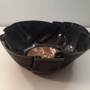 May include: A black bowl crafted from a repurposed vinyl record. The bowl has a unique, wavy shape with cut-out sections. The center of the bowl features the original record label with text. A creative, upcycled home decor item.