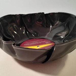 May include: A black vinyl record has been repurposed into a bowl. The record label is visible in the bottom of the bowl. The bowl has a wavy, ruffled edge.