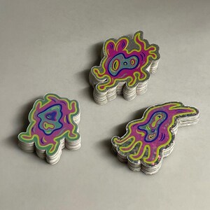 May include: Three stacks of stickers with abstract designs. Each sticker features a unique, colorful illustration with a pink, blue, and green color scheme. The stickers have a silver border and are stacked on a light gray surface.