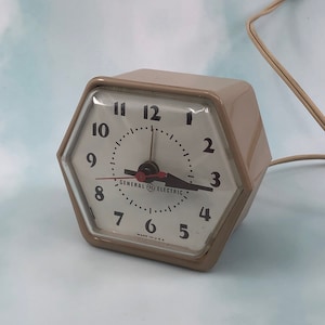 Puede incluir: Un reloj despertador hexagonal beige con números y manecillas negros. La esfera del reloj dice "General Electric" y "Made in U.S.A."