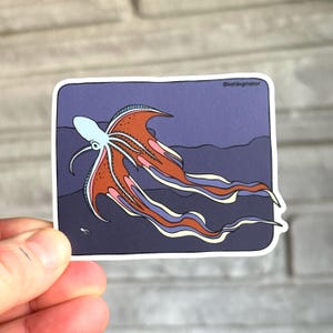 Op de afbeelding: Een sticker met een illustratie van een octopus. De octopus heeft een lichtblauw lichaam en oranje en roze tentakels, tegen een donkerblauwe achtergrond. De tekst "@katdogmanor" staat in de rechterbovenhoek.