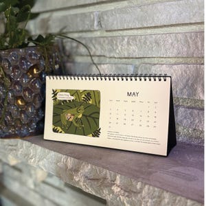 Puede incluir: Un calendario de escritorio para mayo con una ilustración verde de una rana de cristal sobre una hoja. El calendario tiene una base negra y una encuadernación en espiral. La palabra "MAYO" está impresa en la parte superior.