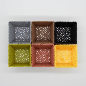 May include: Six square woven baskets in various colors: gray, brown, black, green, red-orange, and yellow. Each basket has a speckled pattern inside. The baskets are arranged in a 2x3 grid on a white background.