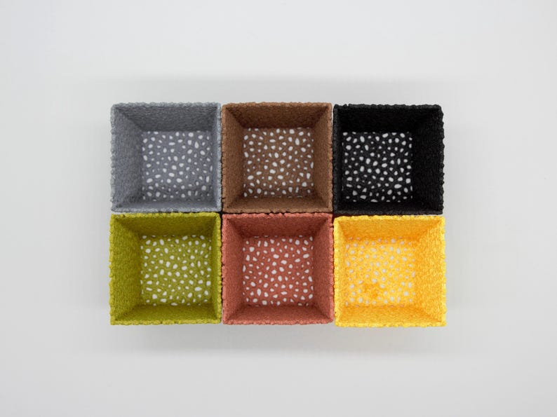 May include: Six square woven baskets in various colors: gray, brown, black, green, red-orange, and yellow. Each basket has a speckled pattern inside. The baskets are arranged in a 2x3 grid on a white background.