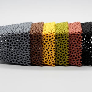 May include: A row of six crocheted cube-shaped objects in various colors: gray, brown, yellow, olive green, reddish-brown, and black. Each cube has a perforated, openwork design, creating a textured appearance. The cubes are arranged side by side.