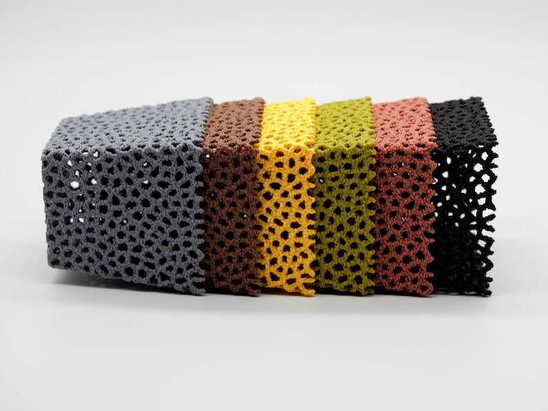 May include: A row of six crocheted cube-shaped objects in various colors: gray, brown, yellow, olive green, reddish-brown, and black. Each cube has a perforated, openwork design, creating a textured appearance. The cubes are arranged side by side.