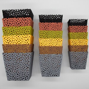 May include: Three sets of colorful, stacked, square planters with a perforated design. The planters are in shades of black, coral, olive green, yellow, brown, and gray. The planters are mounted on a white wall.