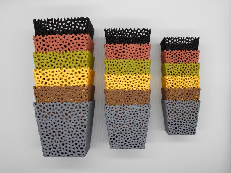 May include: Three sets of colorful, stacked, square planters with a perforated design. The planters are in shades of black, coral, olive green, yellow, brown, and gray. The planters are mounted on a white wall.