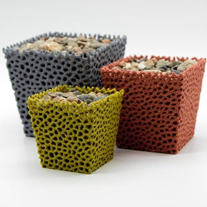 Può includere: Tre fioriere quadrate nei toni del grigio, ruggine e giallo-verde, riempite di piccoli sassi. Le fioriere hanno un design a trama aperta e sono disposte su uno sfondo bianco. Le fioriere sono di dimensioni variabili.