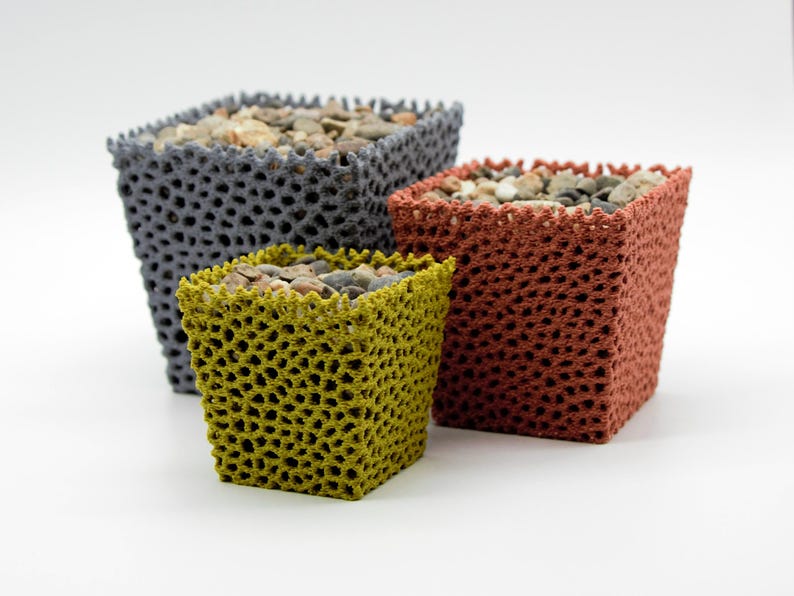 May include: Three square planters in shades of gray, rust, and yellow-green, filled with small stones. The planters have a textured, open-weave design, and are arranged against a white background. The planters are of varying sizes.