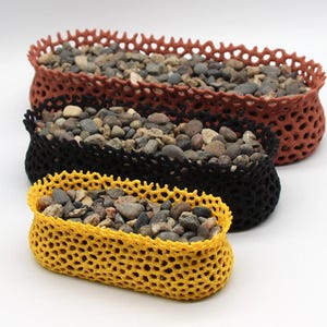 Op de afbeelding: Drie decoratieve manden met open weefsel in verschillende maten en kleuren: roest, zwart en geel. Elke mand is gevuld met kleine, gladde stenen. De manden zijn in een gestapelde formatie gerangschikt en tonen hun unieke texturen en ontwerpen.