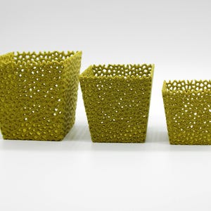 May include: Three olive-green, square-shaped baskets of varying sizes. The baskets have a textured, open-weave design with small holes throughout. The largest basket is approximately 7 inches tall, the medium is 5 inches, and the smallest is 3 inches.