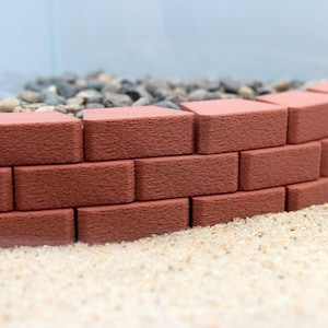 Può includere: Un bordo di mattoni curvo fatto di mattoni rossi, posto su un letto di piccoli ciottoli bianchi. I mattoni sono disposti a spina di pesce, creando un bordo decorativo per un giardino o un vialetto.