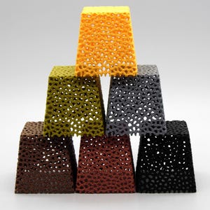 May include: A pyramid of seven openwork, square-based objects in various colors: yellow, olive green, gray, brown, rust, and black. The objects have a honeycomb-like pattern, creating a textured appearance. The objects are stacked on top of each other.
