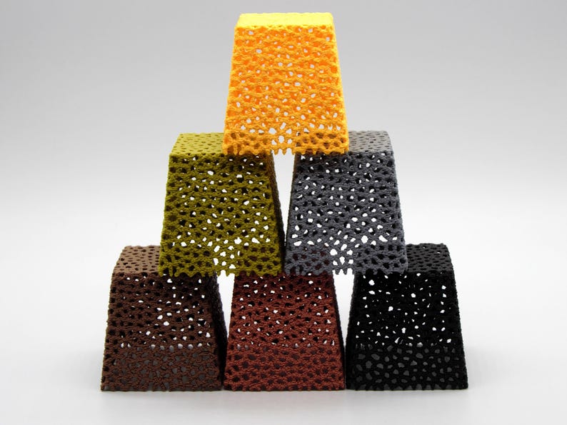 May include: A pyramid of seven openwork, square-based objects in various colors: yellow, olive green, gray, brown, rust, and black. The objects have a honeycomb-like pattern, creating a textured appearance. The objects are stacked on top of each other.