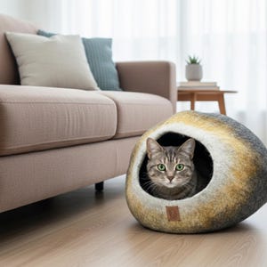 May include: A gray tabby cat peeks out from a round, felted cat bed. The bed has a natural, stone-like appearance with shades of gray, yellow, and brown. A brown leather tag is attached to the bed. A beige sofa and pillows are in the background.