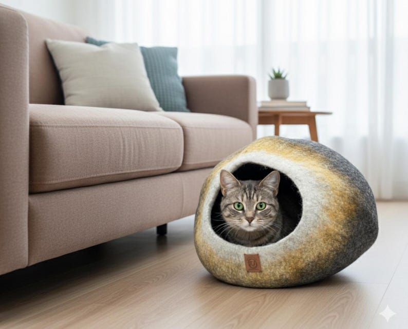 May include: A gray tabby cat peeks out from a round, felted cat bed. The bed has a natural, stone-like appearance with shades of gray, yellow, and brown. A brown leather tag is attached to the bed. A beige sofa and pillows are in the background.