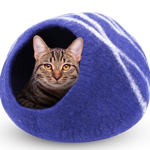 May include: A tabby cat peeks out from a blue cat bed. The bed is a rounded shape with white stripes and a dark blue exterior. The cat has brown and black stripes and yellow eyes.