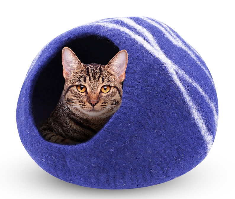 May include: A tabby cat peeks out from a blue cat bed. The bed is a rounded shape with white stripes and a dark blue exterior. The cat has brown and black stripes and yellow eyes.