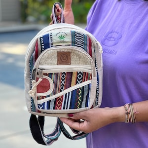 Może przedstawiać: Mały, beżowy plecak z konopi z kolorowymi akcentami z tkanej tkaniny. Plecak posiada przednią kieszeń zapinaną na zamek, górny uchwyt i regulowane paski. Widoczny jest napis "Himalayan Hemp".