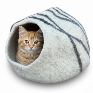 May include: A small orange tabby cat peeks out from a gray and white striped cat bed. The bed is a rounded, cave-like structure made of felted material, providing a cozy and enclosed space for the cat to rest.