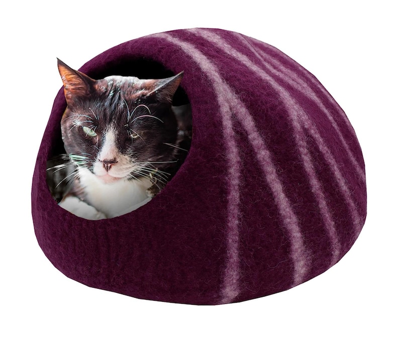 May include: A dark purple cat bed with white stripes, shaped like a dome. A black and white cat is inside the bed, looking out. The bed is made of a soft, felt-like material, providing a cozy space for a pet.