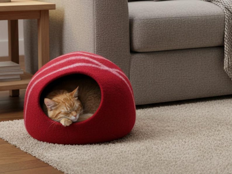 May include: A red cat bed with white stripes, shaped like a dome, sits on a neutral-colored rug. An orange tabby cat is curled up inside the bed, sleeping peacefully. The bed is near a wooden side table and a gray sofa.